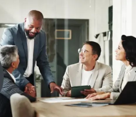 Four businesspeople collaborate about Clariti’s online portal at a table in an office with a laptop, tablet, and paper