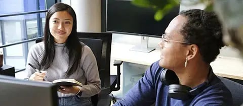Businesswoman with a pen and notebook and businessman with glasses and headphones collaborate at a desk with computers 
