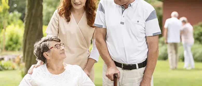An employee at a community living facility assists an elderly lady in a wheel chair and her husband with a walking stick.
