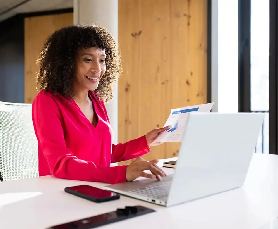 Smiling lady with dark hair at a desk with paper in her hands, while looking at a laptop