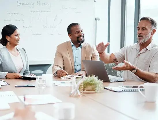 A businessman gestures at a computer screen while two smiling colleagues watch at a brainstorming strategy meeting
