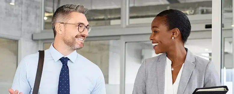 Businesspeople chatting in an office, with one holding a notebook and smartphone and the other carrying a shoulder bag