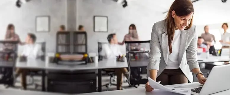 Woman holding papers looks at a laptop screen with her coworkers in the background in a busy office building