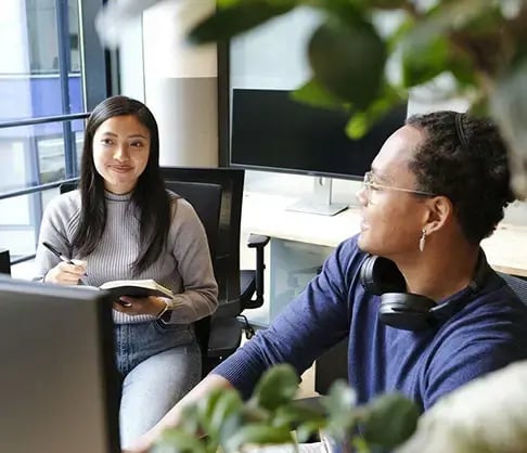 Businesswoman with a pen and notebook and businessman with glasses and headphones collaborate at a desk with computers 